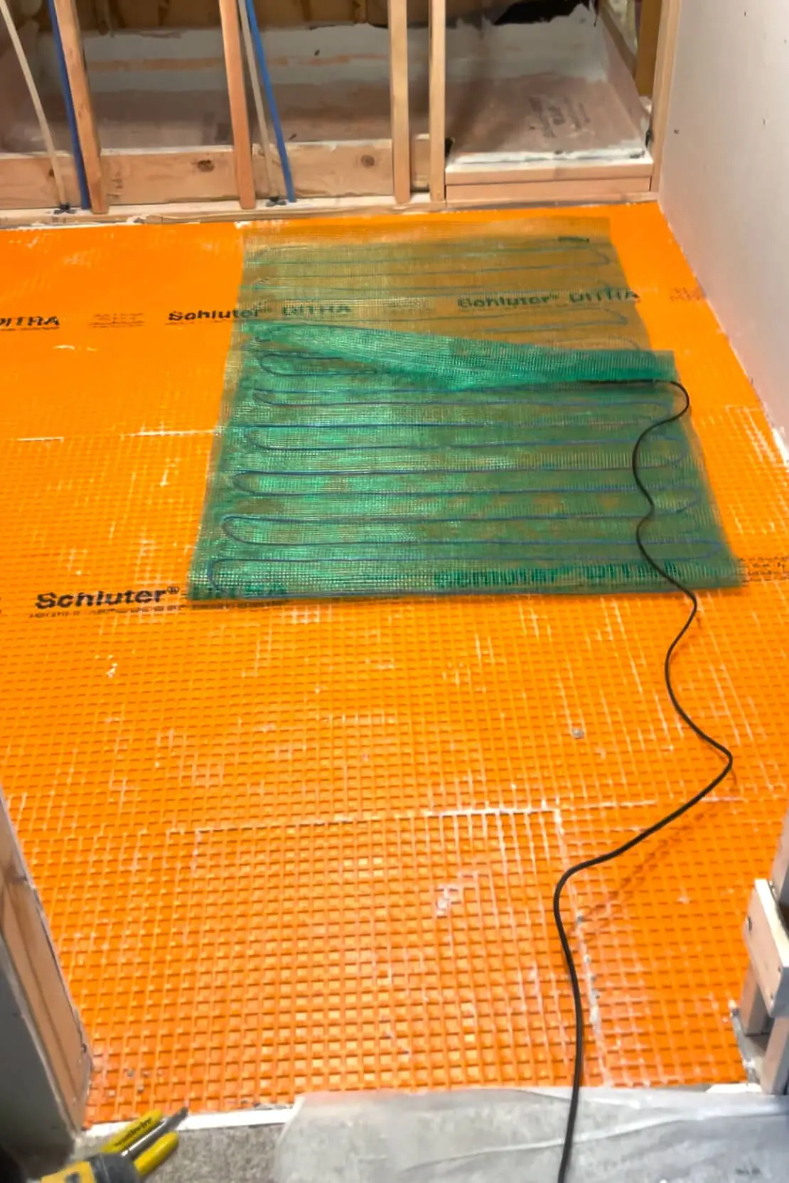 An orange Schluter-DITRA underlayment with a green radiant heating mat laid over it, showing the initial setup stage for a heated bathroom floor system during a renovation.